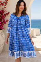 A woman wearing a blue and white patterned bohemian-style dress, standing in front of a Mediterranean-style stone wall with a sea view in the background from Bohemian Love Runway