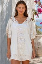 Cream crochet boho kaftan with ornate lace panels and relaxed sleeves in a sunlit Mediterranean coastal setting.