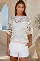 A woman modeling a cream-colored, short-sleeved crochet lace top paired with white shorts, set against a warm beige outdoor architectural background.