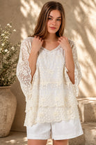 A woman wearing a cream lace top with three-quarter sleeves and white shorts, standing in front of a warm beige wall with leaf shadows.