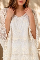 A woman wearing a cream lace top with three-quarter sleeves and white shorts, standing in front of a warm beige wall with leaf shadows.