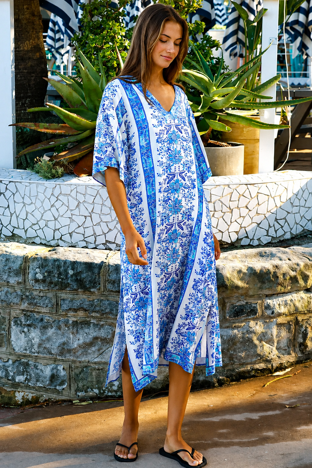 A woman wearing a blue and white patterned kaftan dress with a V-neck and three-quarter sleeves, walking outdoors in front of a stone wall and tropical plants.