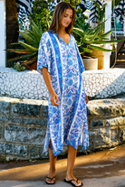 A woman wearing a blue and white patterned kaftan dress with a V-neck and three-quarter sleeves, walking outdoors in front of a stone wall and tropical plants.