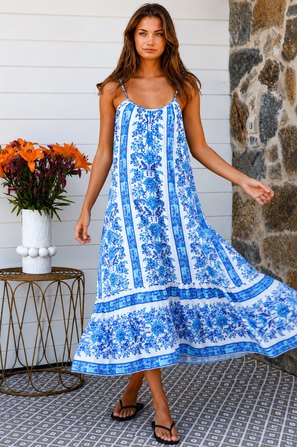 A woman modeling a blue and white patterned maxi dress with spaghetti straps, standing on a patterned tile porch next to a small table with orange flowers.