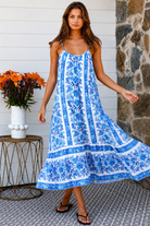 A woman modeling a blue and white patterned maxi dress with spaghetti straps, standing on a patterned tile porch next to a small table with orange flowers.