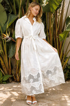 A woman wearing a white lace midi shirt dress with short puff sleeves, a tie waist, and a front slit, standing outdoors in front of tropical green foliage.
