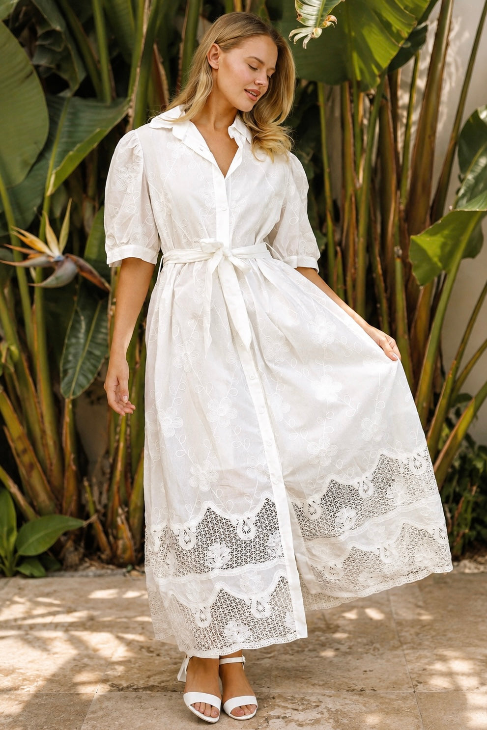 A woman wearing a white lace midi shirt dress with short puff sleeves, a tie waist, and a front slit, standing outdoors in front of tropical green foliage.