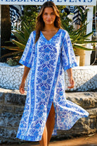 A woman wearing a white kaftan-style tunic dress with a vibrant blue floral and geometric pattern, photographed outdoors with green foliage in the background.