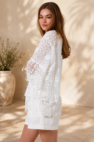 A woman wearing a white lace long-sleeve tunic top with matching white shorts, standing in a sunlit outdoor setting with a beige wall and potted plant in the background.
