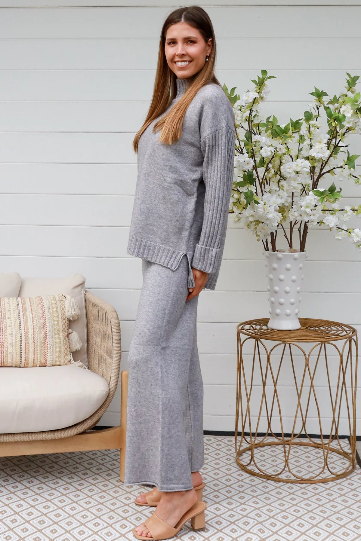 Woman wearing grey knit lounge set with high-neck jumper and wide-leg pants standing in front of a white timber wall - Mellow Lounge Set Grey Mist Bohemian Love Runway 