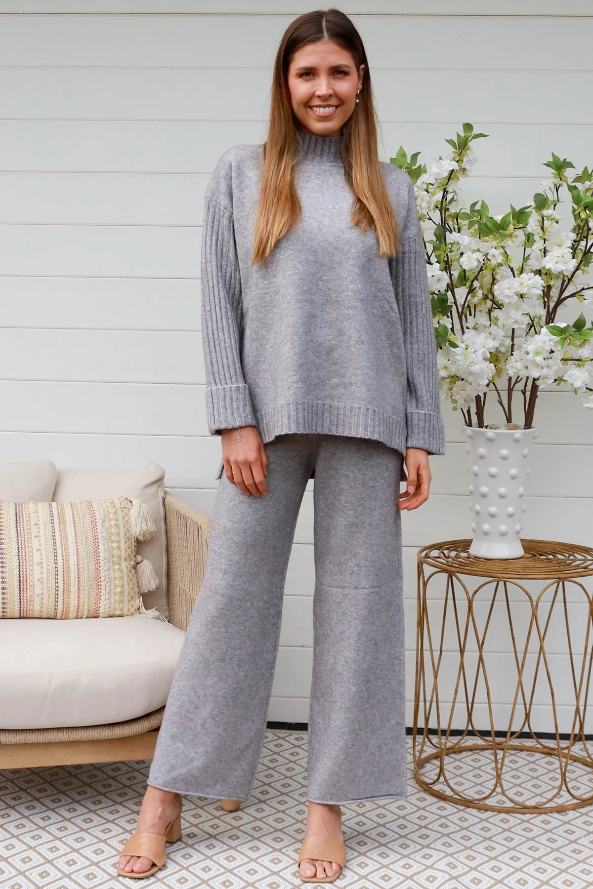 Woman wearing grey knit lounge set with high-neck jumper and wide-leg pants standing in front of a white timber wall - Mellow Lounge Set Grey Mist Bohemian Love Runway 
