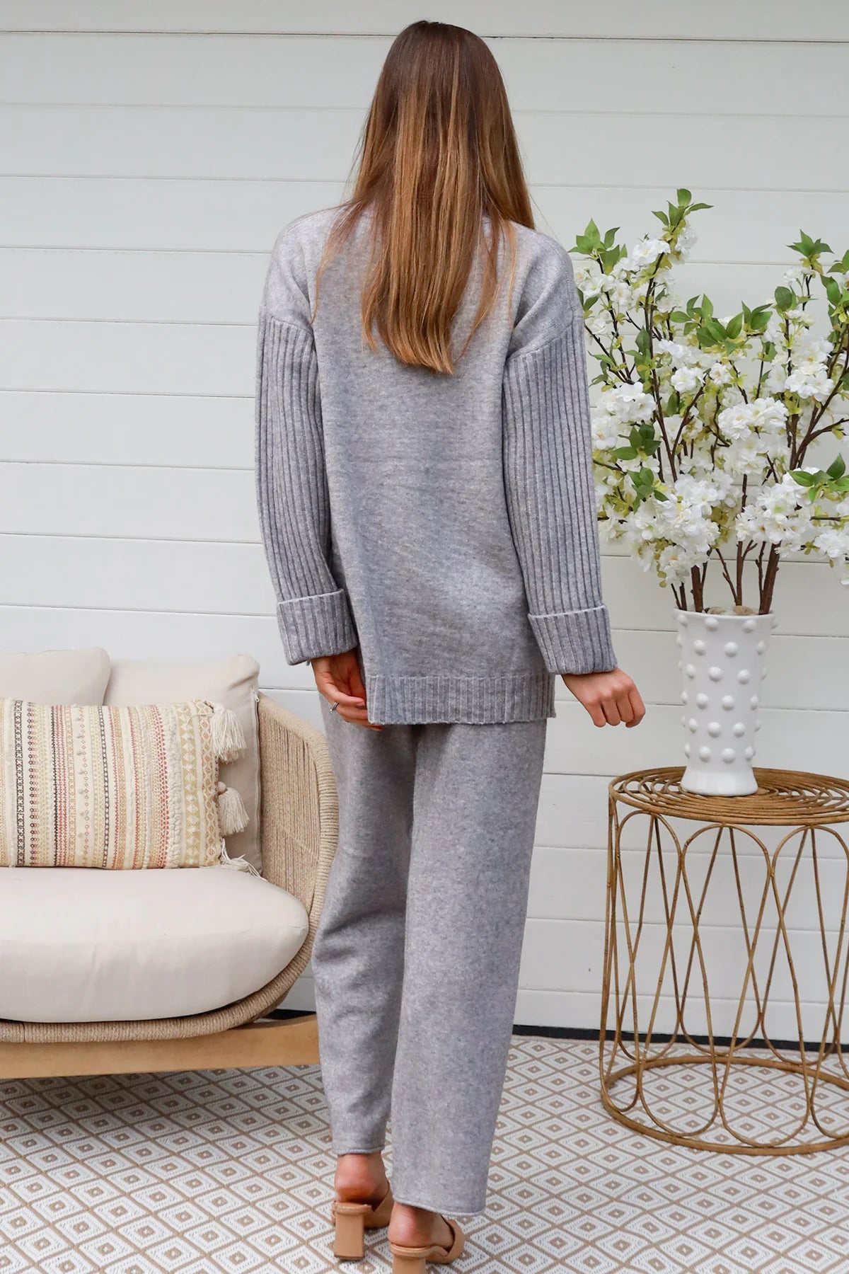 Woman wearing grey knit lounge set with high-neck jumper and wide-leg pants standing in front of a white timber wall - Mellow Lounge Set Grey Mist Bohemian Love Runway 