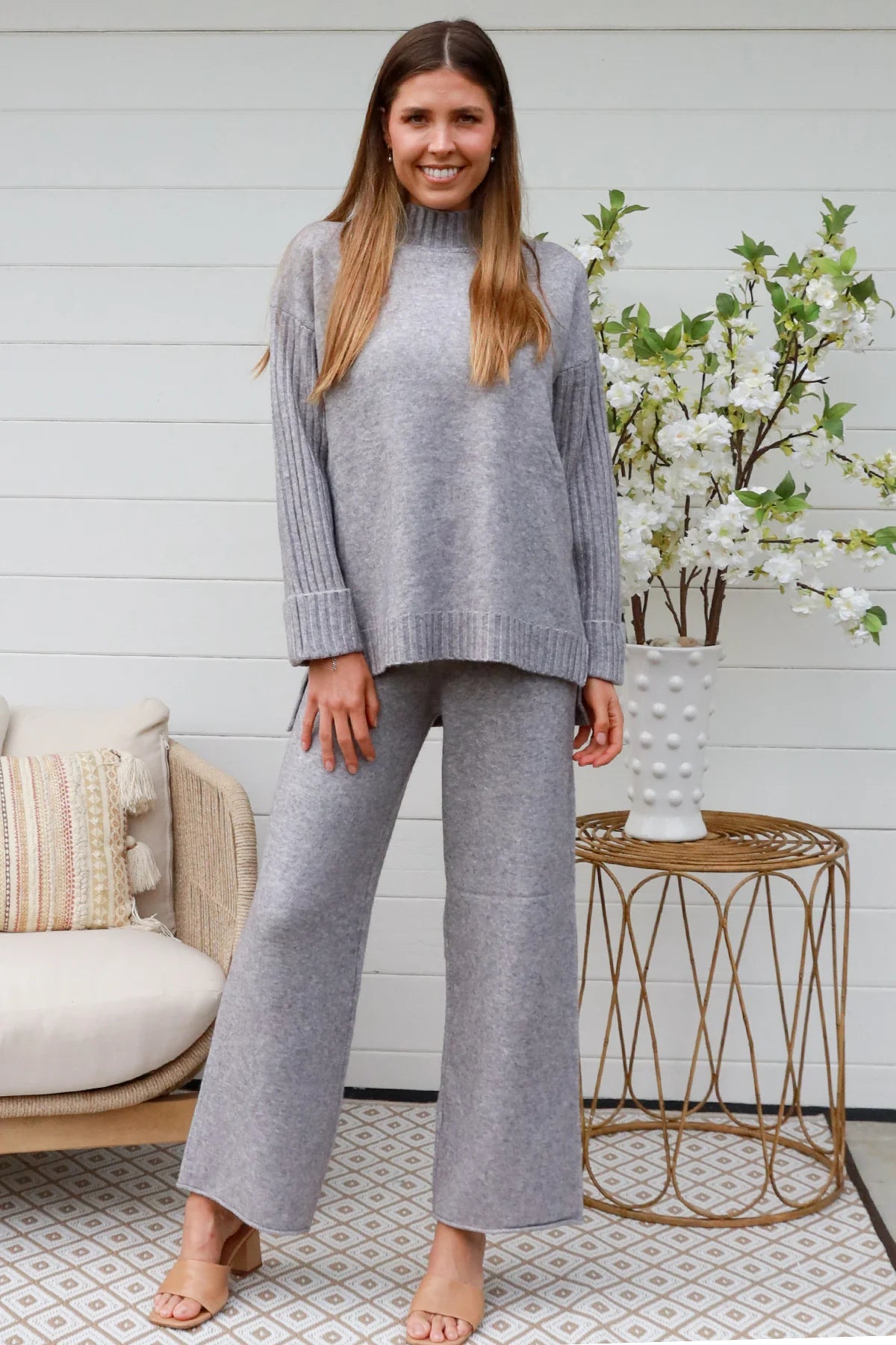 Woman wearing grey knit lounge set with high-neck jumper and wide-leg pants standing in front of a white timber wall - Mellow Lounge Set Grey Mist Bohemian Love Runway 