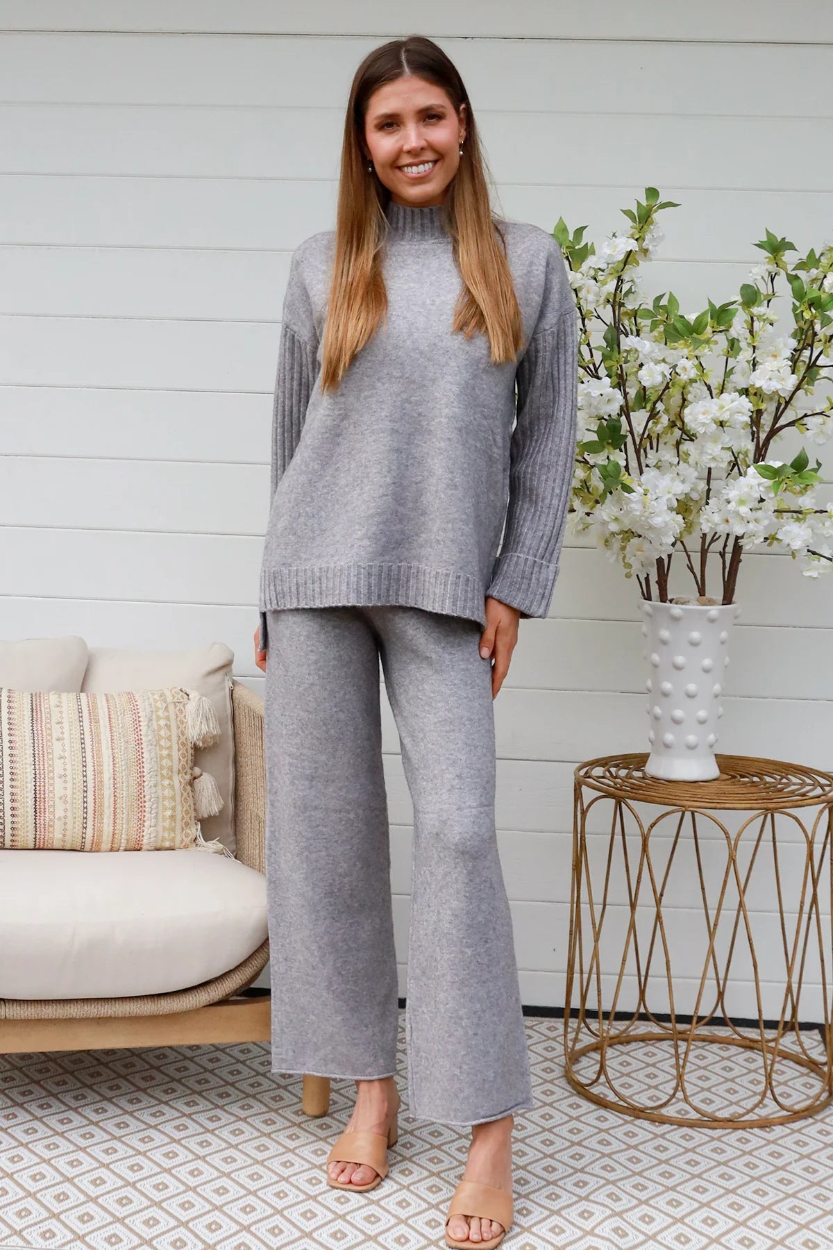 Woman wearing grey knit lounge set with high-neck jumper and wide-leg pants standing in front of a white timber wall - Mellow Lounge Set Grey Mist Bohemian Love Runway 