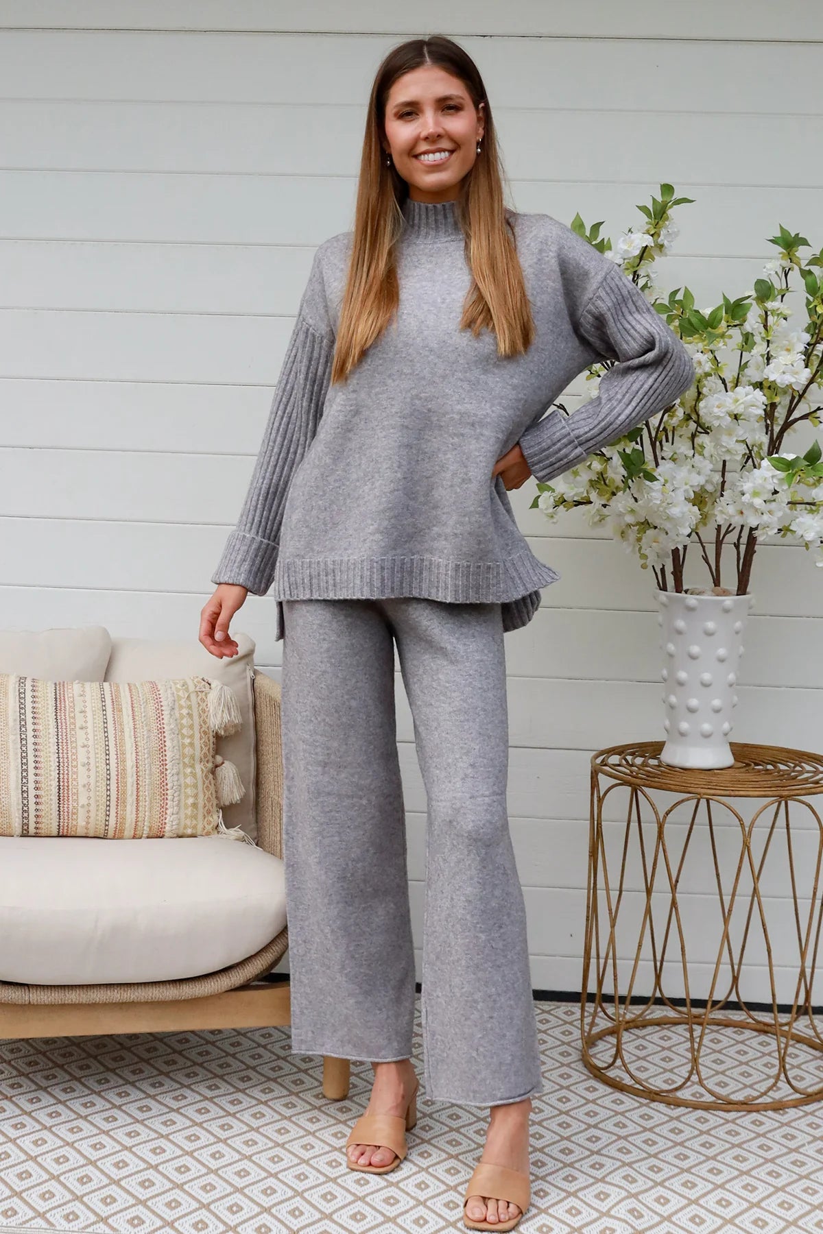 Woman wearing grey knit lounge set with high-neck jumper and wide-leg pants standing in front of a white timber wall - Mellow Lounge Set Grey Mist Bohemian Love Runway 