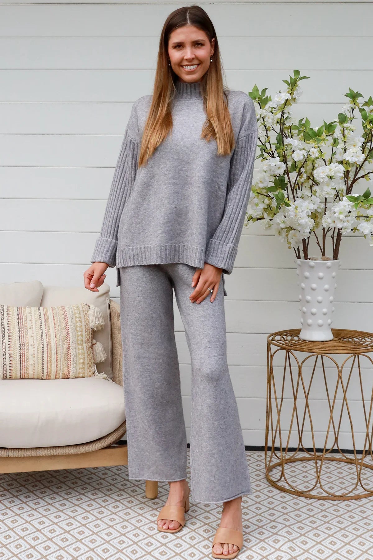 Woman wearing grey knit lounge set with high-neck jumper and wide-leg pants standing in front of a white timber wall - Mellow Lounge Set Grey Mist Bohemian Love Runway 