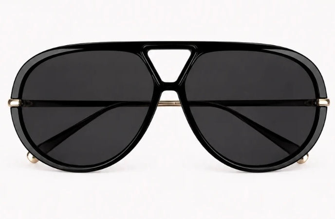 A close-up of a woman wearing oversized black aviator-style sunglasses with gold metal temples, along with gold hoop earrings and gold rings, against a light gray background.