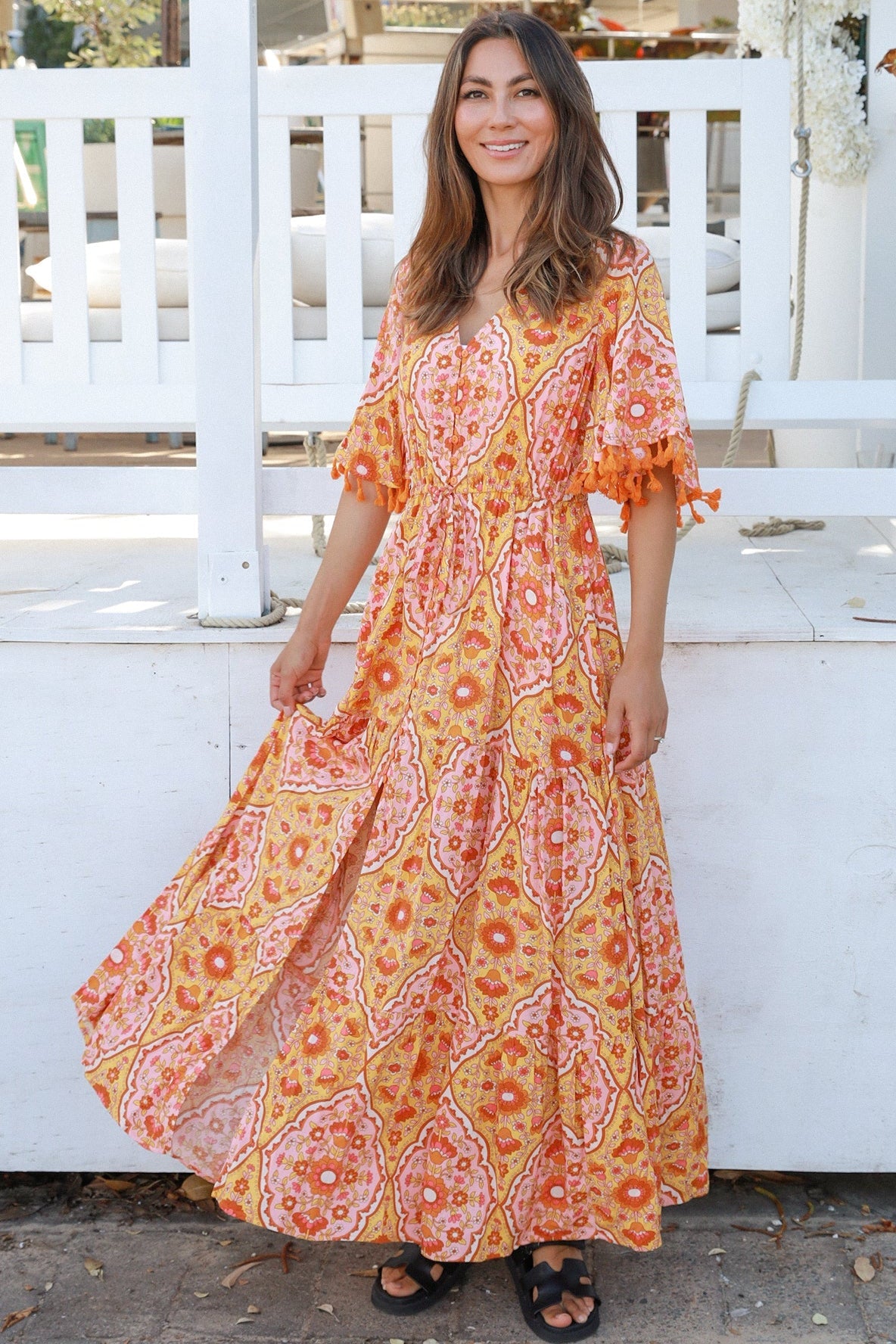 Woman wearing a vintage-inspired boho maxi dress in warm coral and apricot floral print, featuring flutter sleeves, a drawstring waist, and a flowing tiered skirt, styled outdoor in soft summer light.