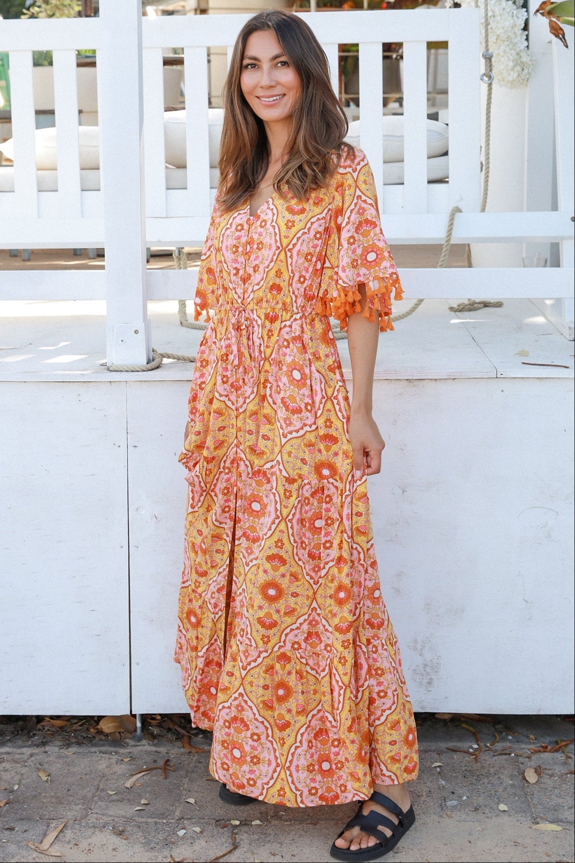 Woman wearing a vintage-inspired boho maxi dress in warm coral and apricot floral print, featuring flutter sleeves, a drawstring waist, and a flowing tiered skirt, styled outdoor in soft summer light.