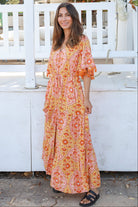 Woman wearing a vintage-inspired boho maxi dress in warm coral and apricot floral print, featuring flutter sleeves, a drawstring waist, and a flowing tiered skirt, styled outdoor in soft summer light.