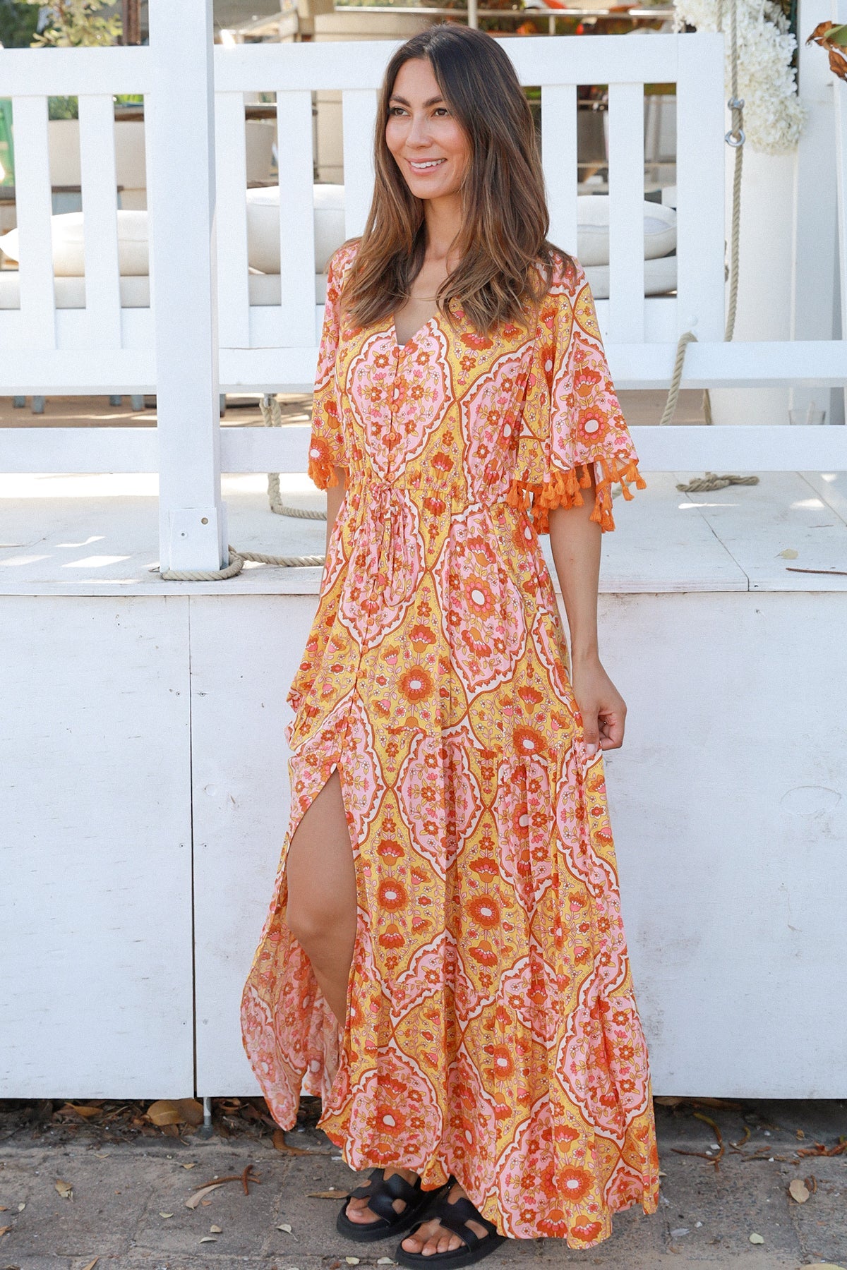 Woman wearing a vintage-inspired boho maxi dress in warm coral and apricot floral print, featuring flutter sleeves, a drawstring waist, and a flowing tiered skirt, styled outdoor in soft summer light.