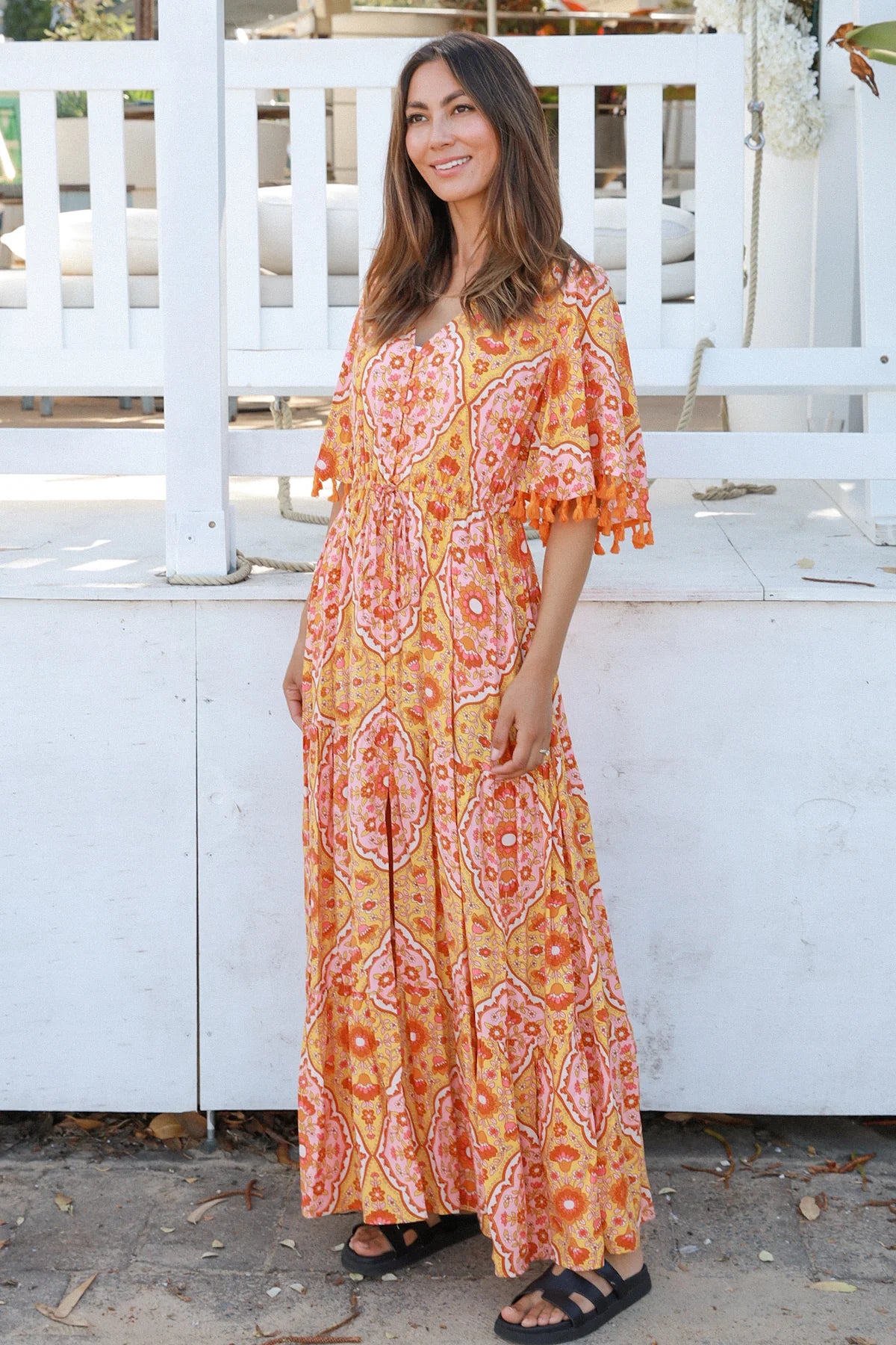 Woman wearing a vintage-inspired boho maxi dress in warm coral and apricot floral print, featuring flutter sleeves, a drawstring waist, and a flowing tiered skirt, styled outdoor in soft summer light.