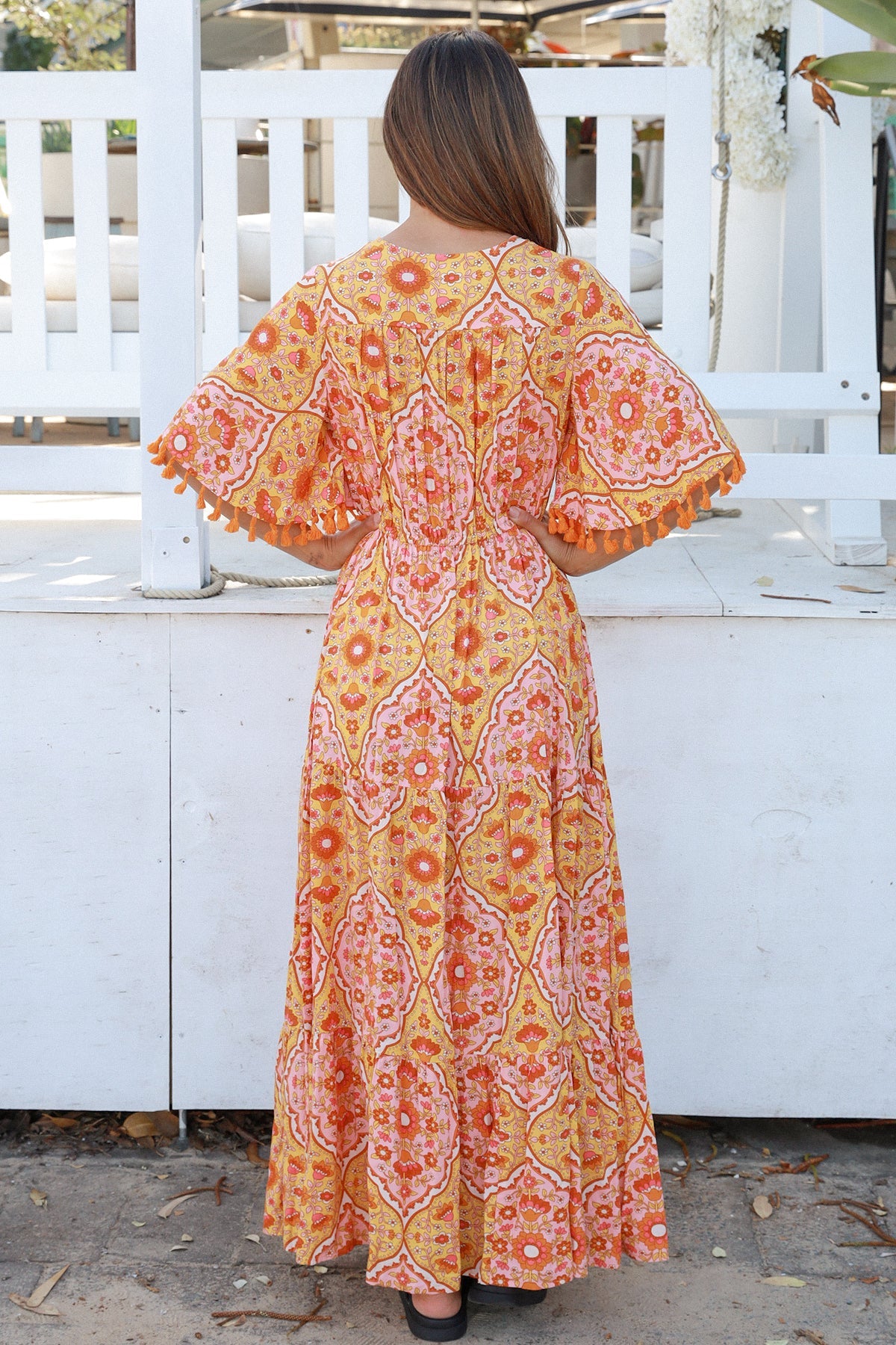 Woman wearing a vintage-inspired boho maxi dress in warm coral and apricot floral print, featuring flutter sleeves, a drawstring waist, and a flowing tiered skirt, styled outdoor in soft summer light.