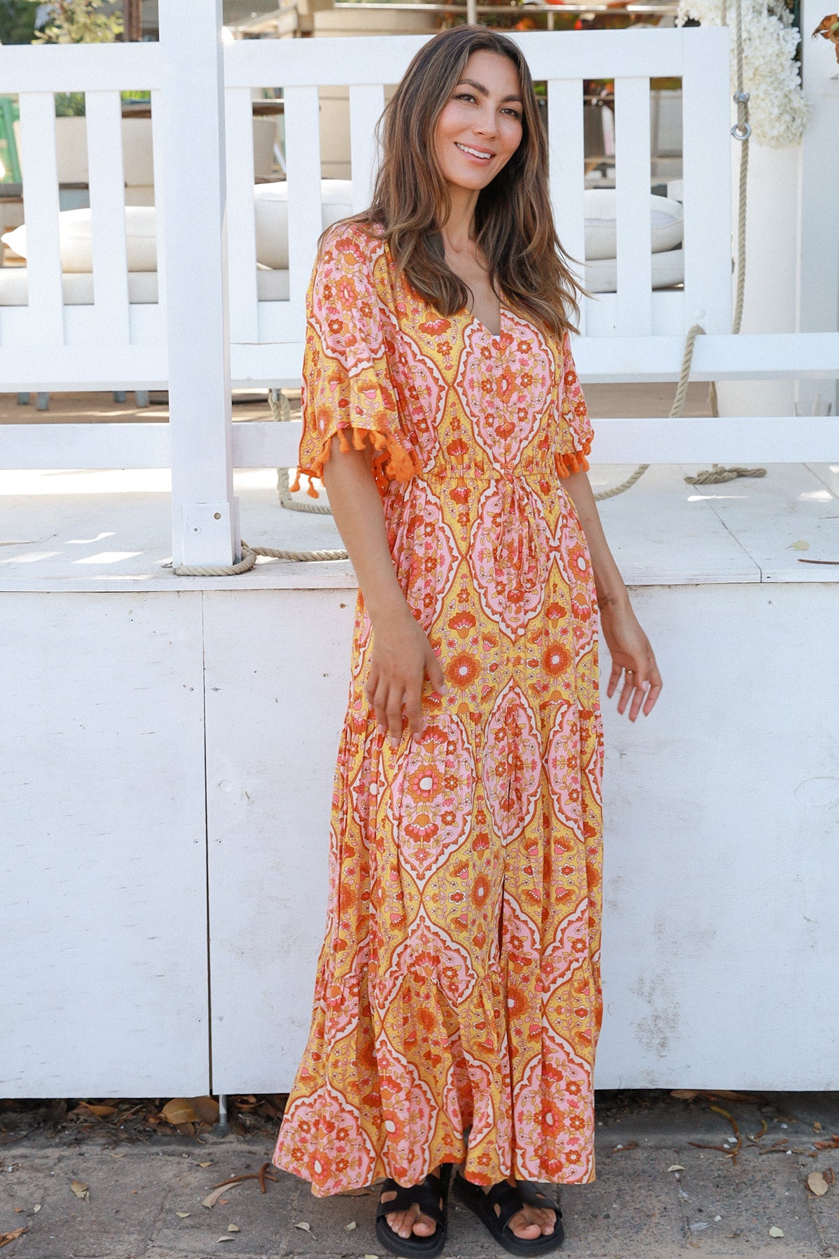 Woman wearing a vintage-inspired boho maxi dress in warm coral and apricot floral print, featuring flutter sleeves, a drawstring waist, and a flowing tiered skirt, styled outdoor in soft summer light.