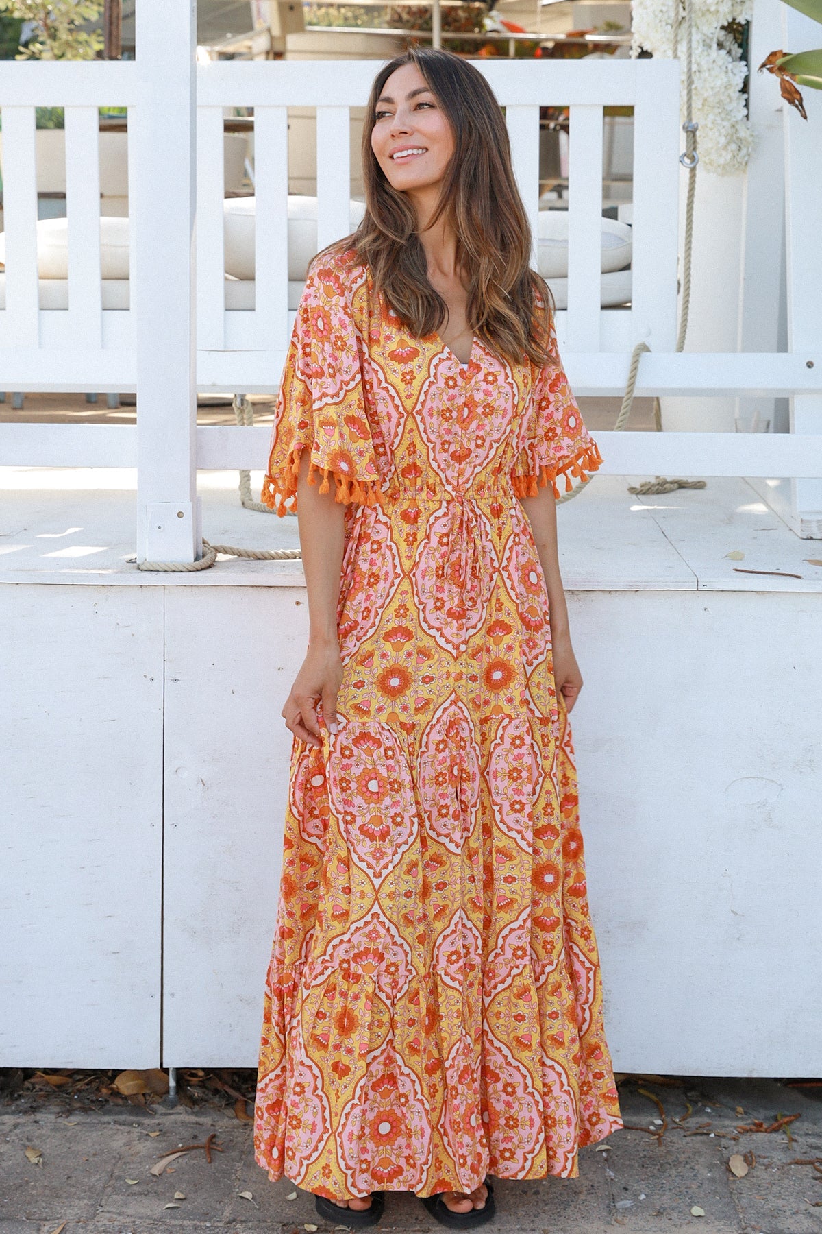 Woman wearing a vintage-inspired boho maxi dress in warm coral and apricot floral print, featuring flutter sleeves, a drawstring waist, and a flowing tiered skirt, styled outdoor in soft summer light.