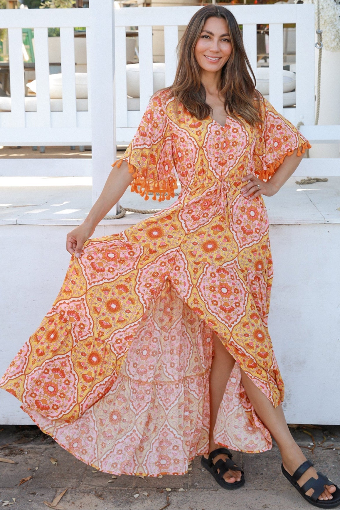 Woman wearing a vintage-inspired boho maxi dress in warm coral and apricot floral print, featuring flutter sleeves, a drawstring waist, and a flowing tiered skirt, styled outdoor in soft summer light.
