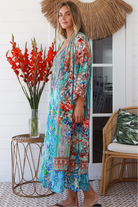 Woman in turquoise floral linen - viscose kimono with bronze and sapphire border detailing, walking along a sunlit coastal setting - resort luxe boho style.