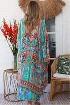 Woman in turquoise floral linen - viscose kimono with bronze and sapphire border detailing, walking along a sunlit coastal setting - resort luxe boho style.