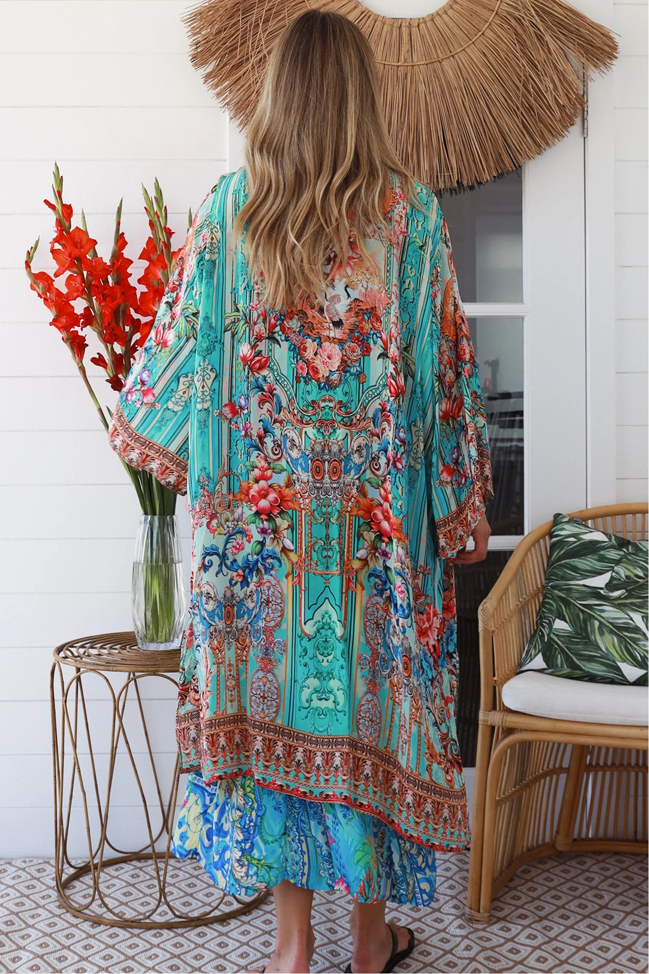 Woman in turquoise floral crepe - viscose kimono with bronze and sapphire border detailing, walking along a sunlit coastal setting - resort luxe boho style.
