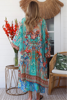 Woman in turquoise floral linen - viscose kimono with bronze and sapphire border detailing, walking along a sunlit coastal setting - resort luxe boho style.