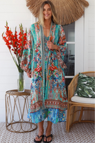 Woman in turquoise floral linen - viscose kimono with bronze and sapphire border detailing, walking along a sunlit coastal setting - resort luxe boho style.