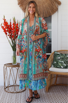 Woman in turquoise floral linen - viscose kimono with bronze and sapphire border detailing, walking along a sunlit coastal setting - resort luxe boho style.