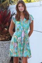Woman wearing a green and pink painterly patchwork rayon mini dress with flutter sleeves and waist tie by Salty Palm