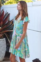 Woman wearing a green and pink painterly patchwork rayon mini dress with flutter sleeves and waist tie by Salty Palm