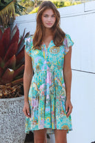 Woman wearing a green and pink painterly patchwork rayon mini dress with flutter sleeves and waist tie by Salty Palm
