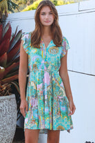 Woman wearing a green and pink painterly patchwork rayon mini dress with flutter sleeves and waist tie by Salty Palm