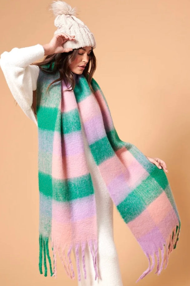 Woman wearing a chunky white knit beanie and oversized fluffy scarf in pastel green, pink and lilac stripes, styled with a neutral winter outfit.