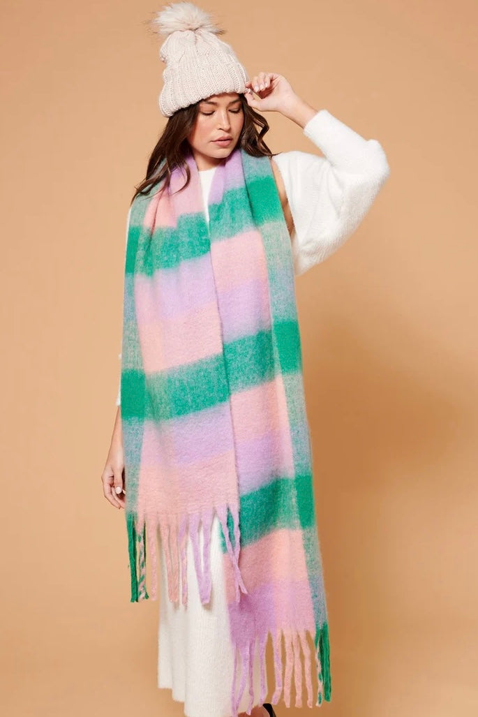 Woman wearing a chunky white knit beanie and oversized fluffy scarf in pastel green, pink and lilac stripes, styled with a neutral winter outfit.