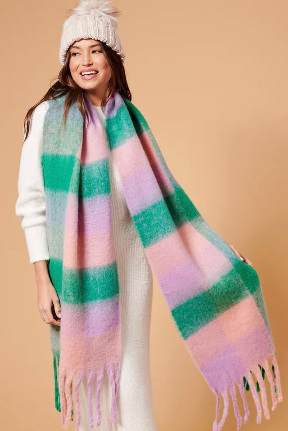 Woman wearing a chunky white knit beanie and oversized fluffy scarf in pastel green, pink and lilac stripes, styled with a neutral winter outfit.