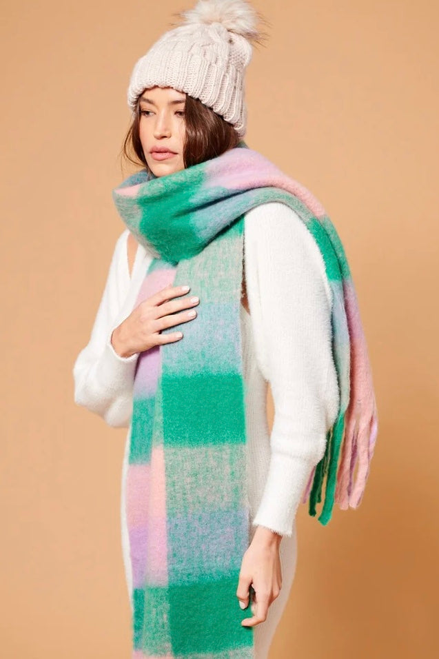 Woman wearing a chunky white knit beanie and oversized fluffy scarf in pastel green, pink and lilac stripes, styled with a neutral winter outfit.