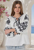 In motion woman wearing a white long-sleeve blouse with black floral embroidery and tie details, paired with light blue jeans, standing in front of a white archway with blue sea and pink flowers in the background from Bohemian Love Runway.