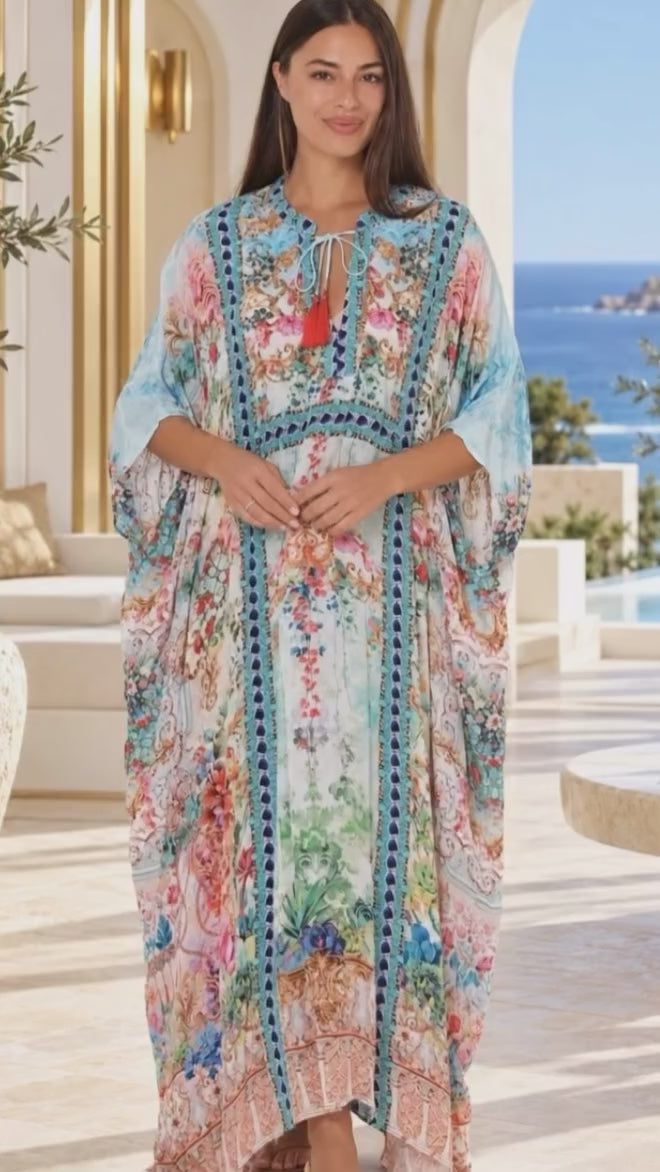 Model in motion wearing a long, flowing kaftan dress with a colorful floral and botanical print adorned with sparkling rhinestones standing on a sunlit outdoor patio with a view of the ocean in the background | Bohemian Love Runway 
