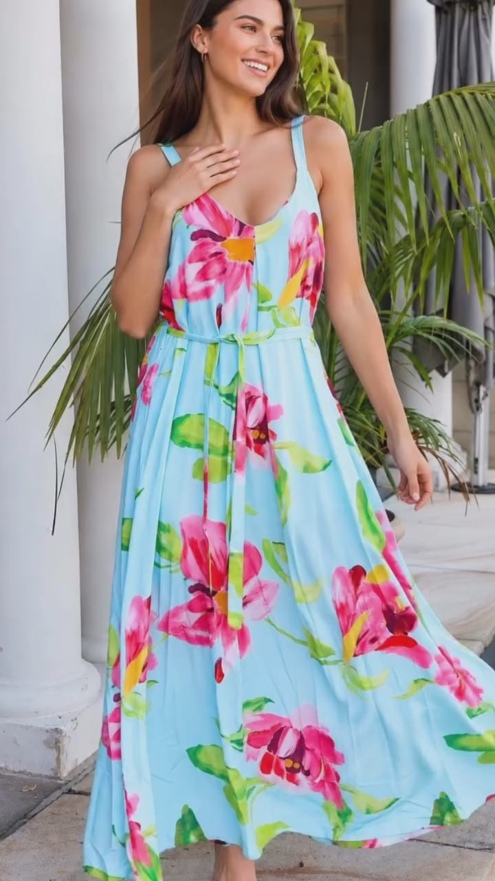Model in motion wearing a light blue sleeveless maxi dress with a vibrant pink and green tropical floral print, standing on stone steps in front of a house with white columns and potted palm trees.