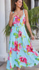 Model in motion wearing a light blue sleeveless maxi dress with a vibrant pink and green tropical floral print, standing on stone steps in front of a house with white columns and potted palm trees.