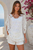In motion from Bohemian Love Runway wearing a white eyelet lace cover-up top with cutout details, standing outdoors in front of a white stucco wall with a blue ocean and pink flowers in the background.