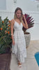 White embroidered cotton maxi dress with lace-trim neckline and scalloped tiered skirt in natural summer light.
